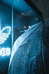 An umbrella covered in raindrops is positioned near a store window, where a bright neon sign featuring a logo is partially visible.