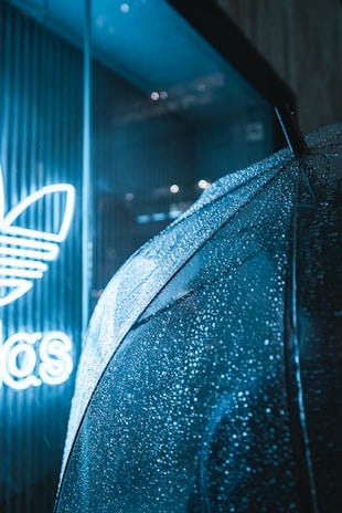 An umbrella covered in raindrops is positioned near a store window, where a bright neon sign featuring a logo is partially visible.