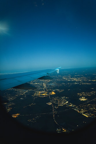 An aerial view during nighttime, capturing the illuminated grid of city lights below with the wing of an airplane prominently in the foreground. The sky above is deep blue, hinting at a clear night.