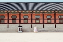 A historical building with traditional architectural features is shown with intricate wooden designs and colorful details along the roof. In front of the building, a couple dressed in traditional clothing stands close together, giving a sense of cultural heritage and romance.