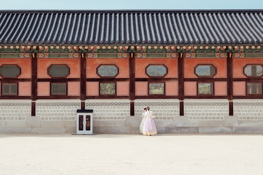 A historical building with traditional architectural features is shown with intricate wooden designs and colorful details along the roof. In front of the building, a couple dressed in traditional clothing stands close together, giving a sense of cultural heritage and romance.