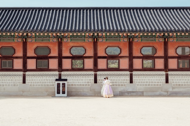 A historical building with traditional architectural features is shown with intricate wooden designs and colorful details along the roof. In front of the building, a couple dressed in traditional clothing stands close together, giving a sense of cultural heritage and romance.
