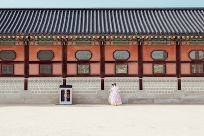 A historical building with traditional architectural features is shown with intricate wooden designs and colorful details along the roof. In front of the building, a couple dressed in traditional clothing stands close together, giving a sense of cultural heritage and romance.
