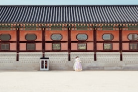 A historical building with traditional architectural features is shown with intricate wooden designs and colorful details along the roof. In front of the building, a couple dressed in traditional clothing stands close together, giving a sense of cultural heritage and romance.
