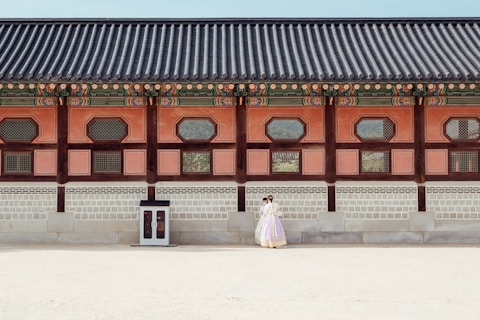 A historical building with traditional architectural features is shown with intricate wooden designs and colorful details along the roof. In front of the building, a couple dressed in traditional clothing stands close together, giving a sense of cultural heritage and romance.