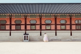 A historical building with traditional architectural features is shown with intricate wooden designs and colorful details along the roof. In front of the building, a couple dressed in traditional clothing stands close together, giving a sense of cultural heritage and romance.