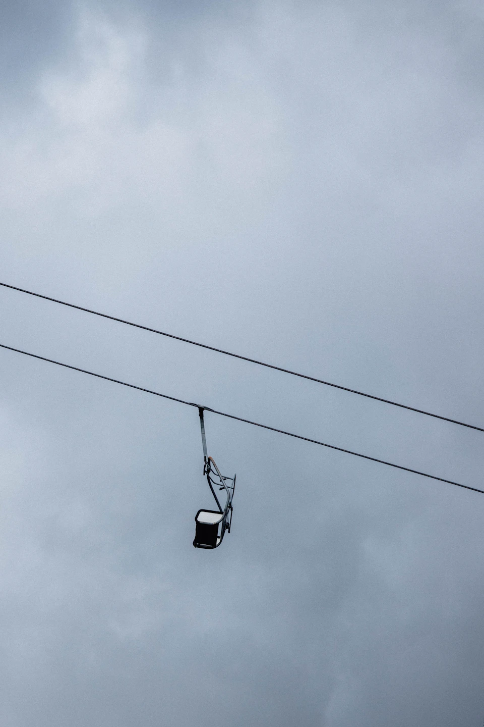 The image depicts a black chair suspended from two wires, positioned at an angle and facing towards the right side of the frame. The sky above is filled with clouds, creating a dramatic backdrop for the solitary chair.
