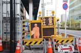 A street scene featuring a construction site with orange traffic cones and a digital sign displaying a construction worker icon and caution symbols in the foreground, set against an urban backdrop with high-rise buildings.