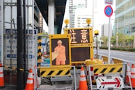 A street scene featuring a construction site with orange traffic cones and a digital sign displaying a construction worker icon and caution symbols in the foreground, set against an urban backdrop with high-rise buildings.