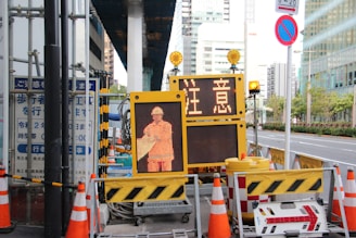 A street scene featuring a construction site with orange traffic cones and a digital sign displaying a construction worker icon and caution symbols in the foreground, set against an urban backdrop with high-rise buildings.