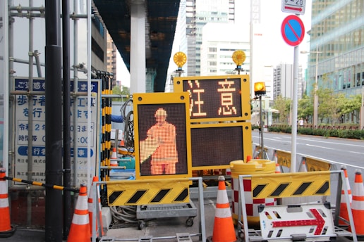A street scene featuring a construction site with orange traffic cones and a digital sign displaying a construction worker icon and caution symbols in the foreground, set against an urban backdrop with high-rise buildings.