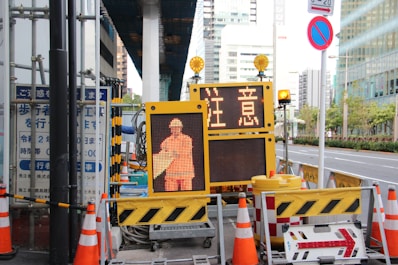 A street scene featuring a construction site with orange traffic cones and a digital sign displaying a construction worker icon and caution symbols in the foreground, set against an urban backdrop with high-rise buildings.