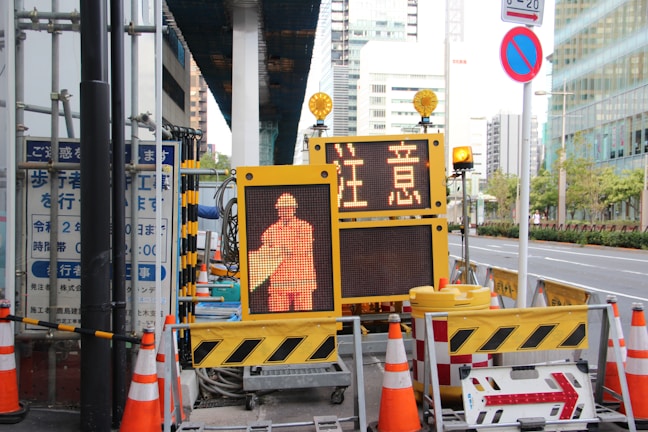 A street scene featuring a construction site with orange traffic cones and a digital sign displaying a construction worker icon and caution symbols in the foreground, set against an urban backdrop with high-rise buildings.