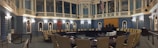 A grand, ornate legislative chamber with blue walls and white columns features a large circular arrangement of desks and chairs. Portraits and statues adorn the upper walls, and flags are displayed prominently. Soft, warm lighting adds a historic atmosphere.