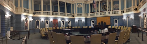 A grand, ornate legislative chamber with blue walls and white columns features a large circular arrangement of desks and chairs. Portraits and statues adorn the upper walls, and flags are displayed prominently. Soft, warm lighting adds a historic atmosphere.
