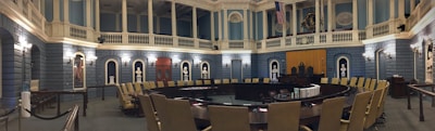 A grand, ornate legislative chamber with blue walls and white columns features a large circular arrangement of desks and chairs. Portraits and statues adorn the upper walls, and flags are displayed prominently. Soft, warm lighting adds a historic atmosphere.