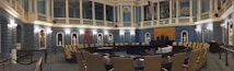 A grand, ornate legislative chamber with blue walls and white columns features a large circular arrangement of desks and chairs. Portraits and statues adorn the upper walls, and flags are displayed prominently. Soft, warm lighting adds a historic atmosphere.