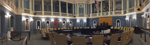 A grand, ornate legislative chamber with blue walls and white columns features a large circular arrangement of desks and chairs. Portraits and statues adorn the upper walls, and flags are displayed prominently. Soft, warm lighting adds a historic atmosphere.