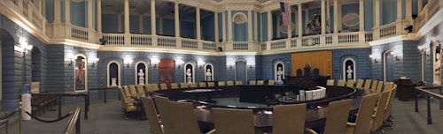 A grand, ornate legislative chamber with blue walls and white columns features a large circular arrangement of desks and chairs. Portraits and statues adorn the upper walls, and flags are displayed prominently. Soft, warm lighting adds a historic atmosphere.