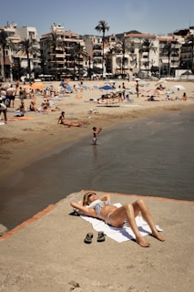 A person relaxes on a sandy beachfront, lying on a towel and wearing sunglasses and a striped swimsuit. The beach is bustling with people enjoying the sun, swimming, and lounging under colorful umbrellas. Palm trees and multi-story buildings line the background, enhancing the coastal atmosphere.