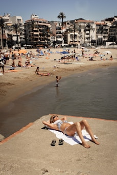 A person relaxes on a sandy beachfront, lying on a towel and wearing sunglasses and a striped swimsuit. The beach is bustling with people enjoying the sun, swimming, and lounging under colorful umbrellas. Palm trees and multi-story buildings line the background, enhancing the coastal atmosphere.