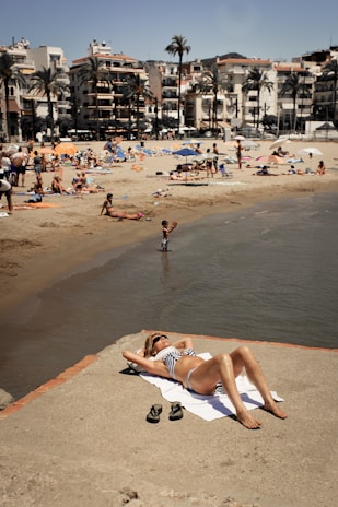 A person relaxes on a sandy beachfront, lying on a towel and wearing sunglasses and a striped swimsuit. The beach is bustling with people enjoying the sun, swimming, and lounging under colorful umbrellas. Palm trees and multi-story buildings line the background, enhancing the coastal atmosphere.