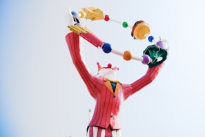 A colorful clown statue with a striped red suit is juggling objects, including a hamburger and other abstract shapes. The clown has a playful pose with arms raised high.