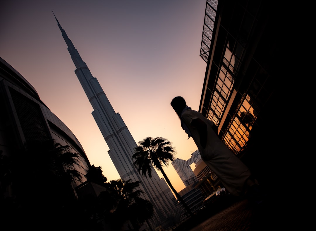 person standing near tower, A low angle shot of a person standing near Burj Khalifa hotel during sunset in Dubai, UAE<p></figure>