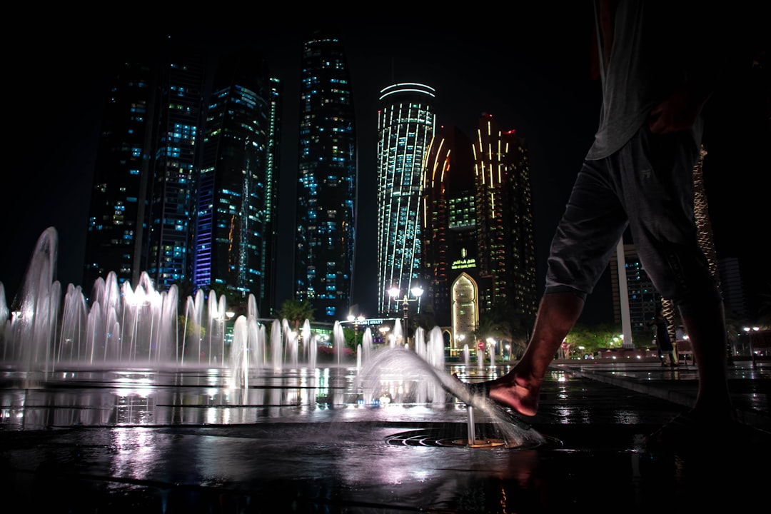 water fountain in front of building during nighttime, A low angle shot of a barefoot person walking on groundwater fountains near Etihad Towers at night<p></figure>