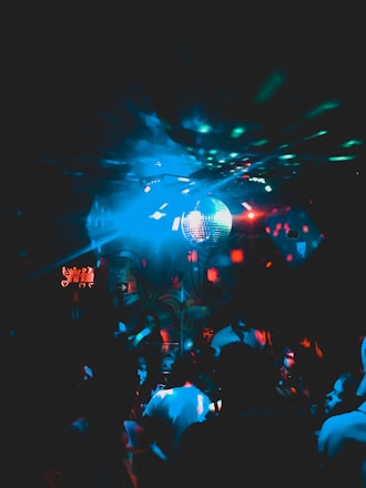 A dimly lit nightclub scene with a crowd of people dancing. A blue disco ball is suspended from the ceiling, casting multicolored light reflections across the room. The background has abstract wall art and small glowing signs.