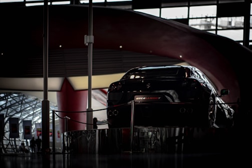 A sleek black sports car is displayed in a modern exhibition space with curved, elegant architecture. Soft lighting illuminates the car from above, highlighting its glossy surface. The background shows a large window with geometric designs, and silhouettes of people are visible nearby.