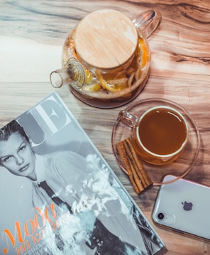 A neatly arranged scene featuring a magazine with a black-and-white cover next to a glass teapot filled with lemon slices and herbs. Beside the teapot is a clear glass cup containing a warm drink, accompanied by two cinnamon sticks. An iPhone with a visible Apple logo rests on the wooden tabletop.