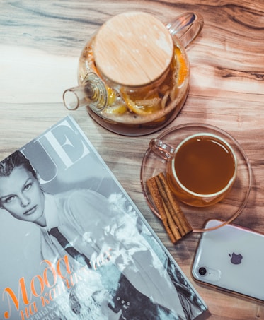 A neatly arranged scene featuring a magazine with a black-and-white cover next to a glass teapot filled with lemon slices and herbs. Beside the teapot is a clear glass cup containing a warm drink, accompanied by two cinnamon sticks. An iPhone with a visible Apple logo rests on the wooden tabletop.