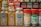 A collection of spice jars and seasoning bottles is arranged on a shelf. The jars include organic curry powder, ground turmeric, ground ginger, lemon pepper seasoning, and parsley flakes. Each jar has distinct labels with branding and certification marks such as USDA Organic and Non-GMO verified. The background shows more spice bottles with various labels and colors, creating a vibrant display typical of a spice rack or kitchen pantry.