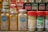 A collection of spice jars and seasoning bottles is arranged on a shelf. The jars include organic curry powder, ground turmeric, ground ginger, lemon pepper seasoning, and parsley flakes. Each jar has distinct labels with branding and certification marks such as USDA Organic and Non-GMO verified. The background shows more spice bottles with various labels and colors, creating a vibrant display typical of a spice rack or kitchen pantry.