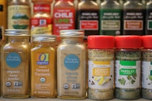 A collection of spice jars and seasoning bottles is arranged on a shelf. The jars include organic curry powder, ground turmeric, ground ginger, lemon pepper seasoning, and parsley flakes. Each jar has distinct labels with branding and certification marks such as USDA Organic and Non-GMO verified. The background shows more spice bottles with various labels and colors, creating a vibrant display typical of a spice rack or kitchen pantry.