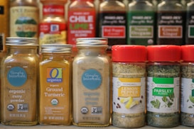 A collection of spice jars and seasoning bottles is arranged on a shelf. The jars include organic curry powder, ground turmeric, ground ginger, lemon pepper seasoning, and parsley flakes. Each jar has distinct labels with branding and certification marks such as USDA Organic and Non-GMO verified. The background shows more spice bottles with various labels and colors, creating a vibrant display typical of a spice rack or kitchen pantry.