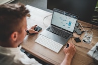 A person is working at a wooden desk with a laptop displaying a calendar or scheduling application. They are using a mouse and a smartphone is nearby, with a glass of water on a coaster and a set of keys on the desk.