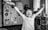 A cheerful child with glasses is raising their arms in excitement. The room is filled with toys, including a toy gun in the child's hand. The background shows a wardrobe with drawings and a stack of board games.