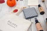 A wooden surface displays a small orange pumpkin, woven items, a patterned cloth, bottles labeled with numbers, and a cream-colored pouch with 'PAIMA' branding. Autumn leaves and natural tones create a cozy, fall-themed setting.