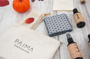 A wooden surface displays a small orange pumpkin, woven items, a patterned cloth, bottles labeled with numbers, and a cream-colored pouch with 'PAIMA' branding. Autumn leaves and natural tones create a cozy, fall-themed setting.