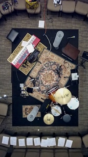 A music setup viewed from above, with a keyboard, drums, microphones, and various audio equipment laid out on intricately patterned rugs. Surrounding the setup are several chairs with sheets of paper scattered around the area.