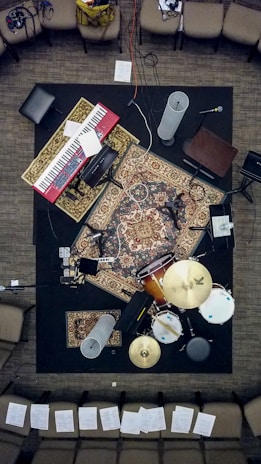 A music setup viewed from above, with a keyboard, drums, microphones, and various audio equipment laid out on intricately patterned rugs. Surrounding the setup are several chairs with sheets of paper scattered around the area.