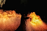 A performer breathes a large burst of fire against a pitch-black background, creating a vivid display of flames and sparks. The contrast between the dark surroundings and the bright fire highlights the dynamic movement and energy of the scene. The performer is wearing protective gear, visible in silhouette.