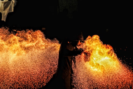 A performer breathes a large burst of fire against a pitch-black background, creating a vivid display of flames and sparks. The contrast between the dark surroundings and the bright fire highlights the dynamic movement and energy of the scene. The performer is wearing protective gear, visible in silhouette.