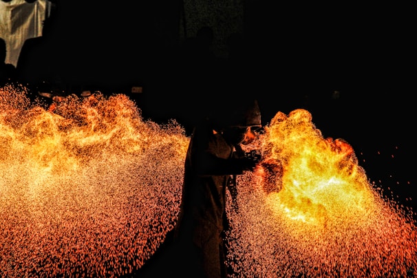 A performer breathes a large burst of fire against a pitch-black background, creating a vivid display of flames and sparks. The contrast between the dark surroundings and the bright fire highlights the dynamic movement and energy of the scene. The performer is wearing protective gear, visible in silhouette.
