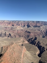 A vast and rugged canyon landscape with layers of multicolored rock formations stretching out to the horizon. The canyon has deep crevices and steep cliffs with various shades of red, brown, and grey. The sky is clear and blue, providing a stark contrast to the earthy tones of the terrain.