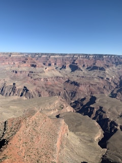 A vast and rugged canyon landscape with layers of multicolored rock formations stretching out to the horizon. The canyon has deep crevices and steep cliffs with various shades of red, brown, and grey. The sky is clear and blue, providing a stark contrast to the earthy tones of the terrain.
