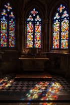 Intricately designed stained glass windows in vibrant reds, blues, and yellows cast colorful light on a checkered stone floor and a small altar. The architectural details and religious imagery create a solemn and reflective atmosphere in the dimly lit space.