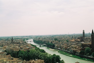 A picturesque urban landscape featuring an expansive view of a historical city with densely packed buildings. A river snakes through the city, bordered by lush greenery. Tall, historical towers and church spires are scattered throughout, standing prominently against the hazy sky.